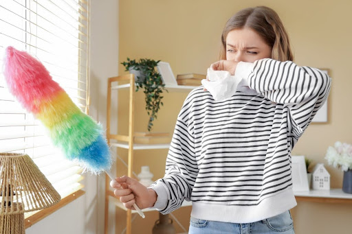 A woman covering her nose and holding a tissue as she dusts window blinds in a home.